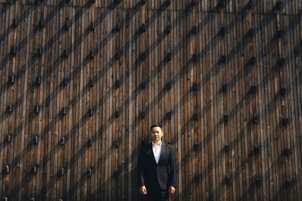 Groom in black suit stands thoughtfully against textured moody wooden wall backdrop.