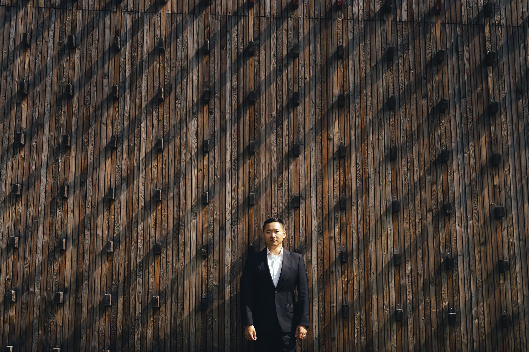 Groom in black suit stands thoughtfully against textured moody wooden wall backdrop.