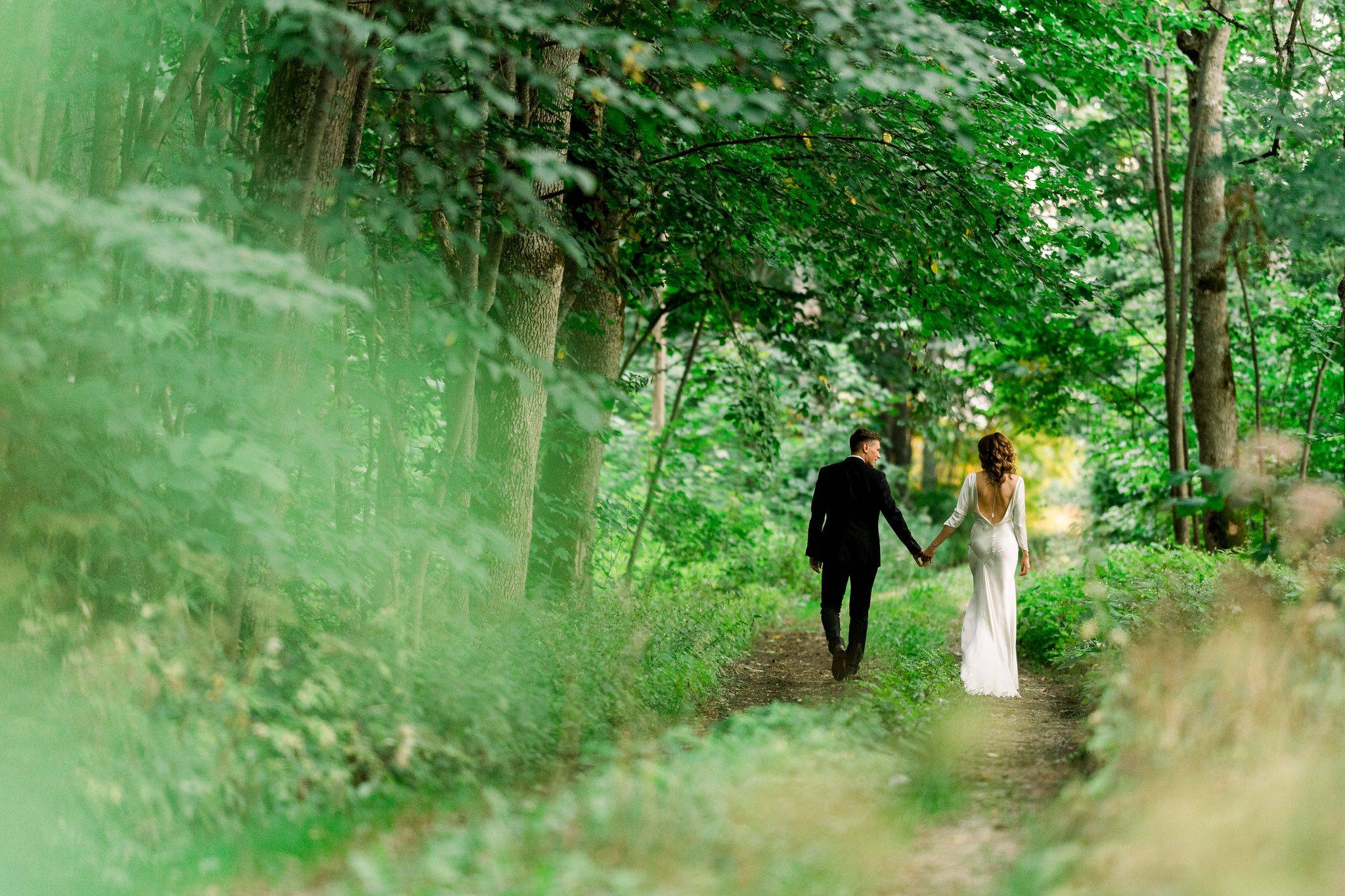 Elegant couple walking hand in hand on a green forest path during wedding photoshoot.