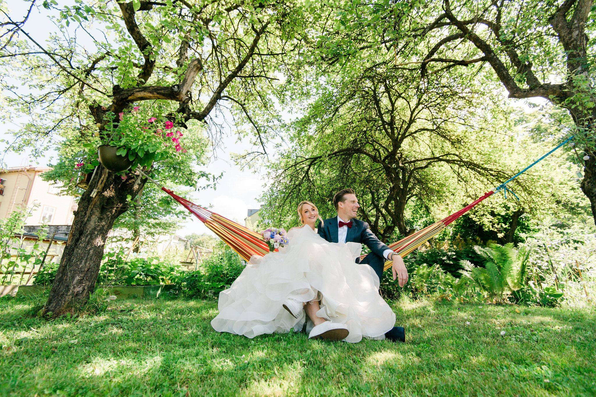 Newlywed couple relaxing in colorful garden hammock surrounded by vibrant greenery and flowers.
