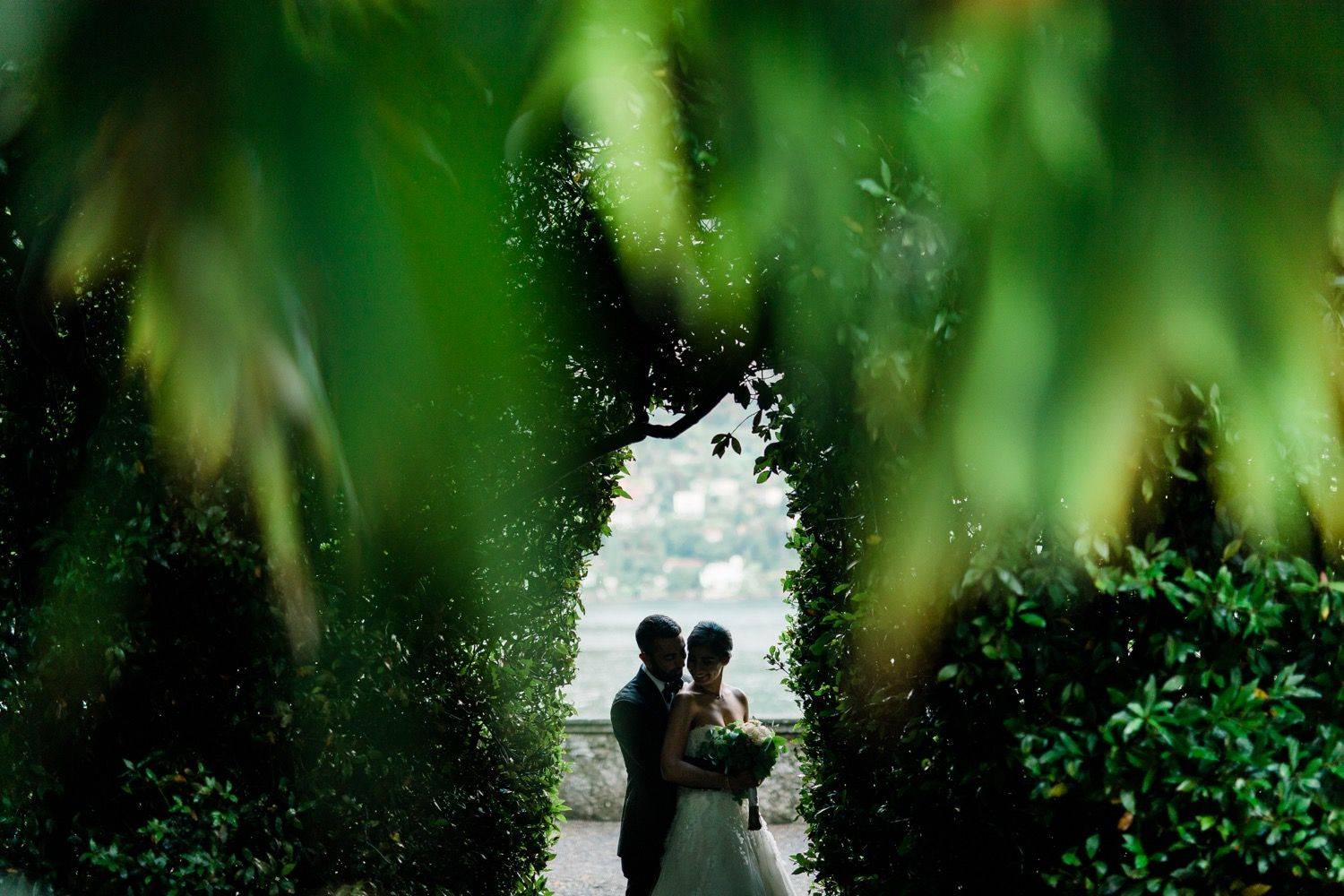 Bride and groom embrace beneath lush green arch with scenic lakeside town background.