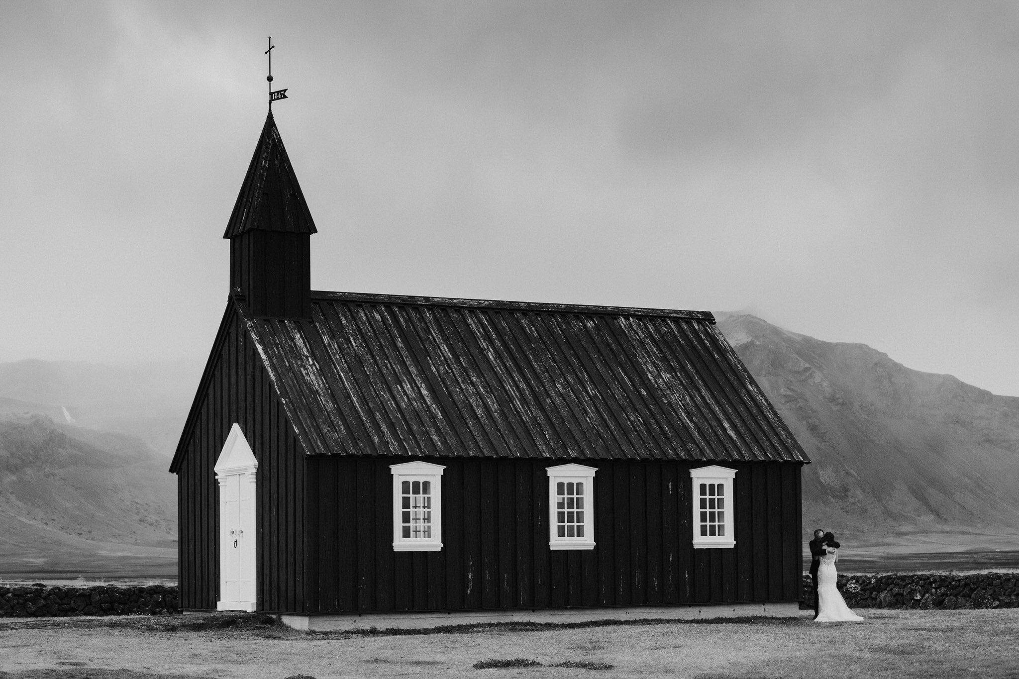 Romantic couple embraces beside iconic black church in remote icelandic mountain landscape.