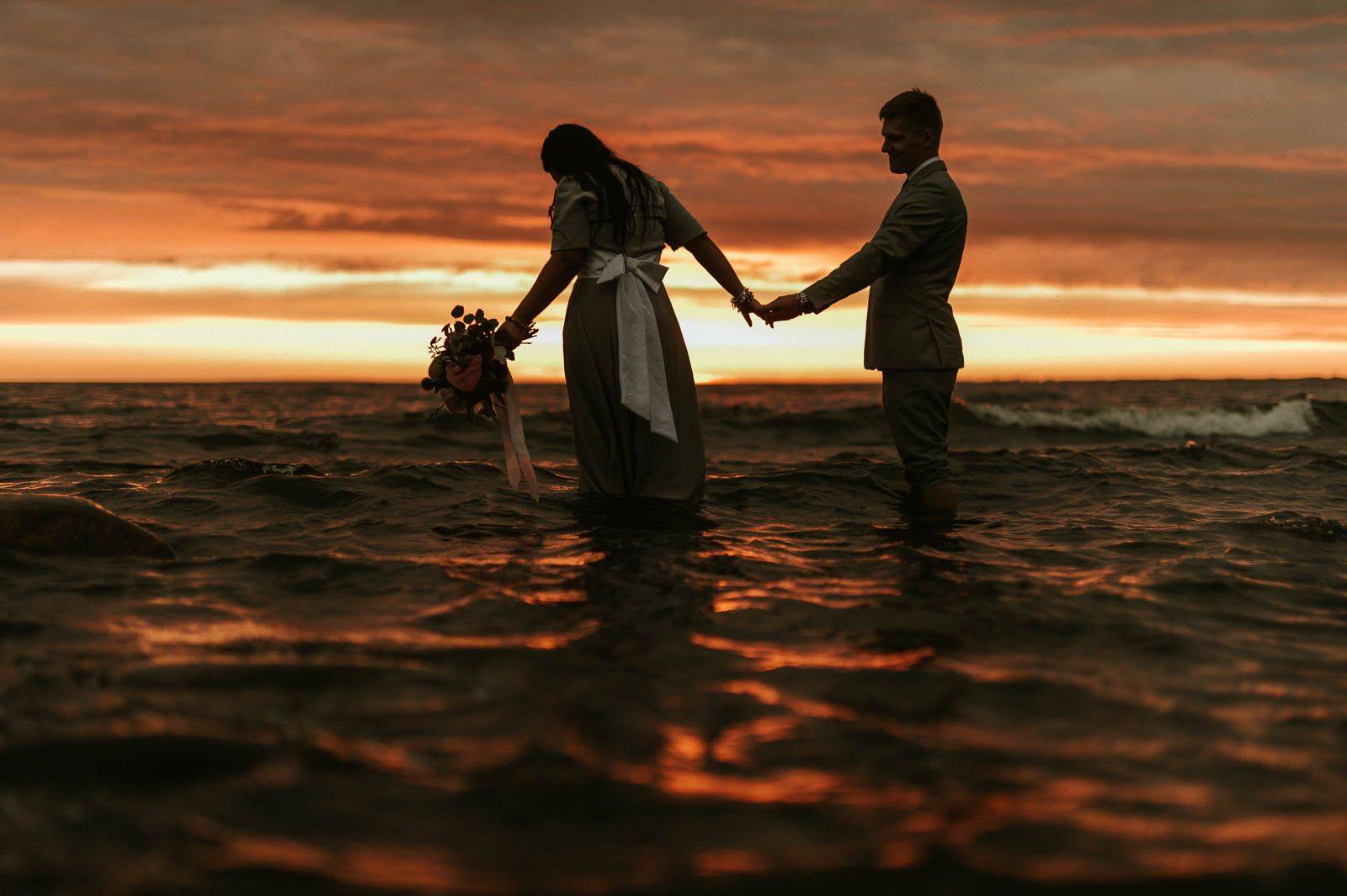Romantic couple holding hands in ocean water during vibrant sunset silhouette.
