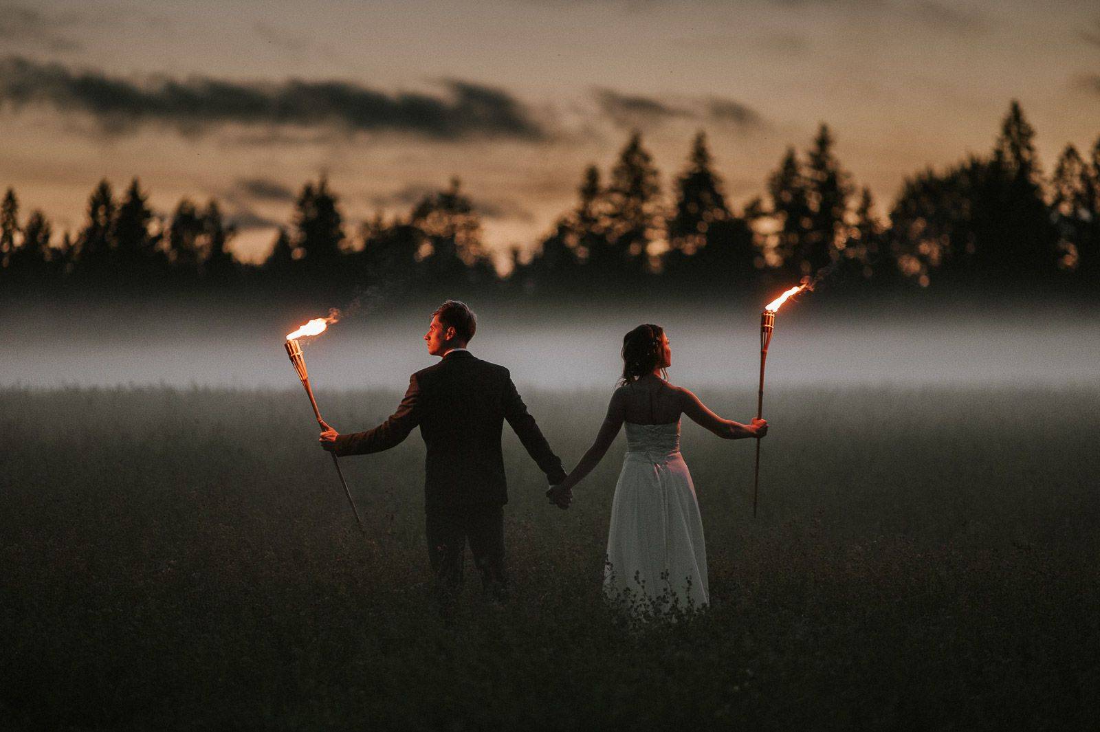 Couple holding torches in misty field at twilight near forest, romantic elopement scene.