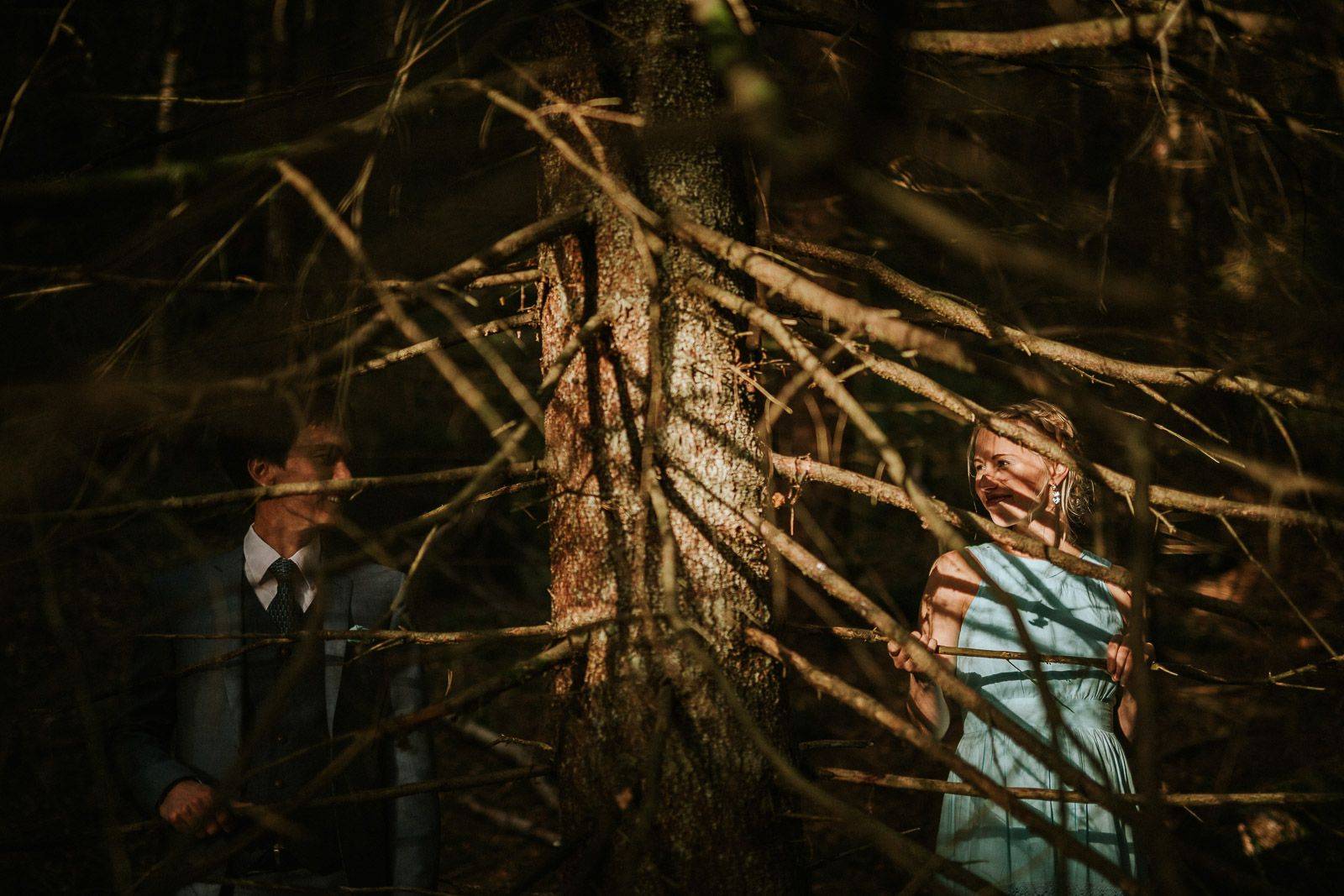 Couple shares a tender moment framed by tree branches in a shadowed icelandic forest.