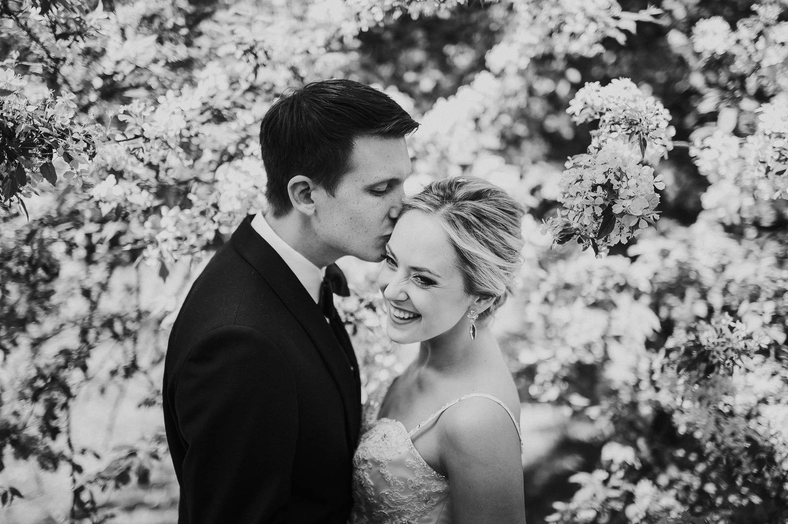 Romantic black-and-white wedding portrait of couple sharing a tender forehead kiss outdoors.