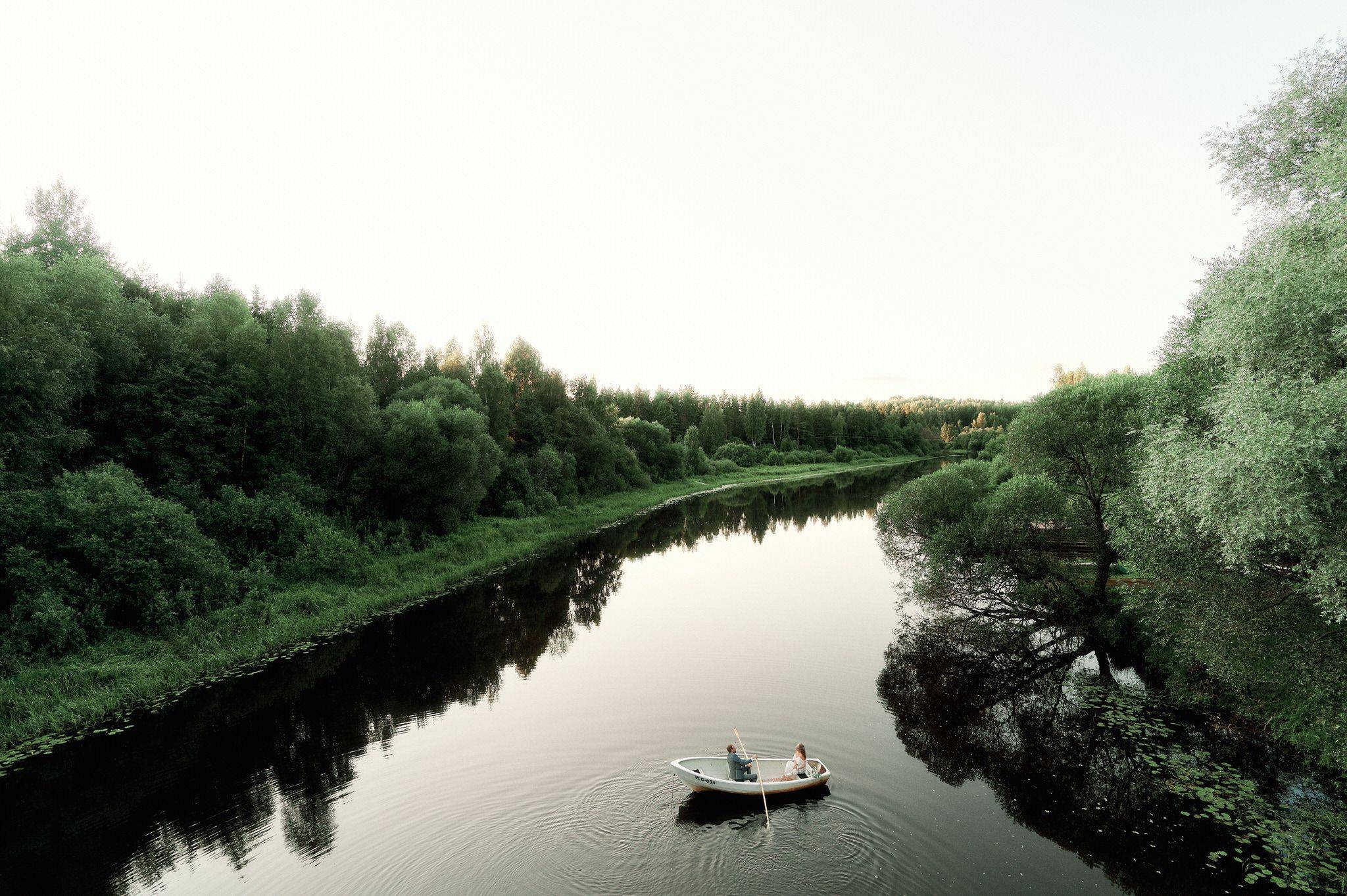 Couple rowing white boat on peaceful river surrounded by lush green trees and calm water.
