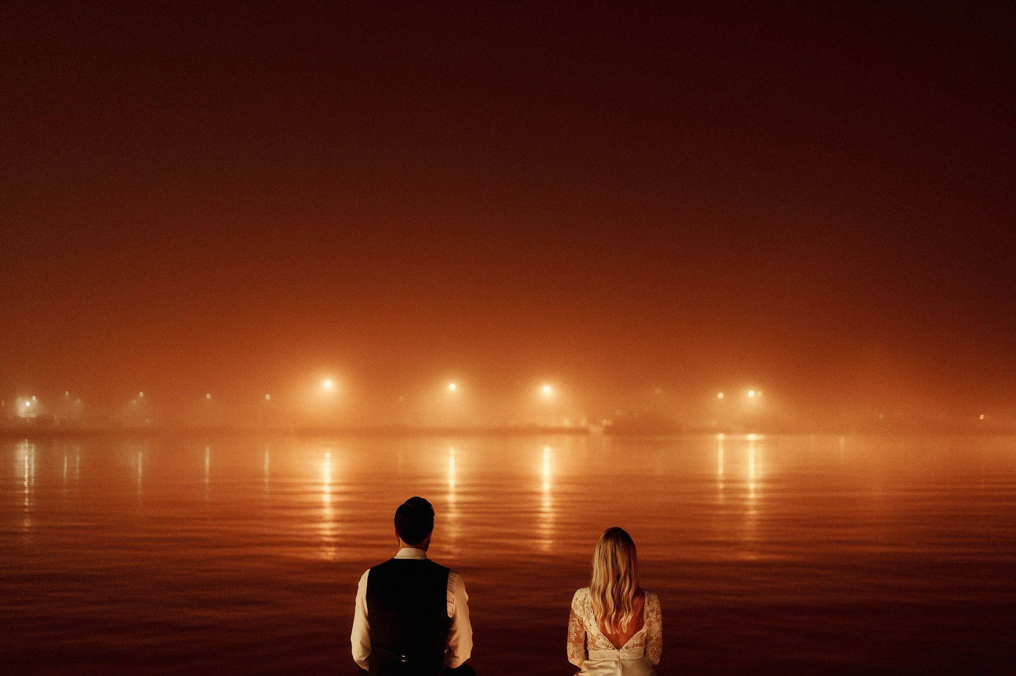 Romantic couple embraces by misty waterfront at night with warm glowing lights reflection.