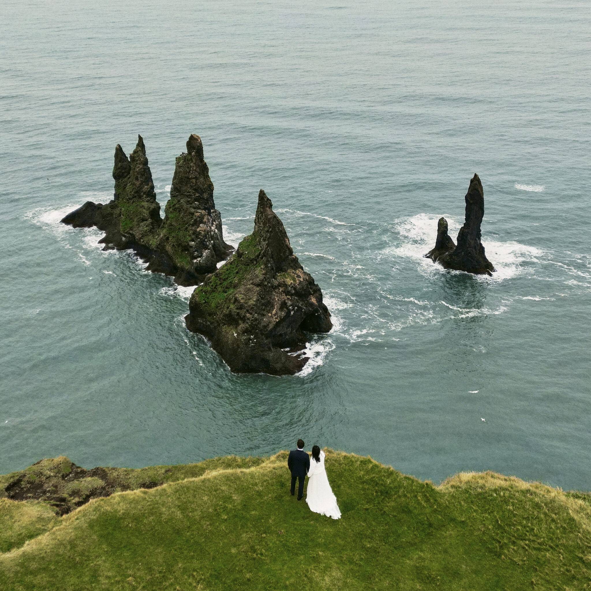 Romantic couple holding hands on green iceland cliffs overlooking jagged sea rocks and calm ocean.