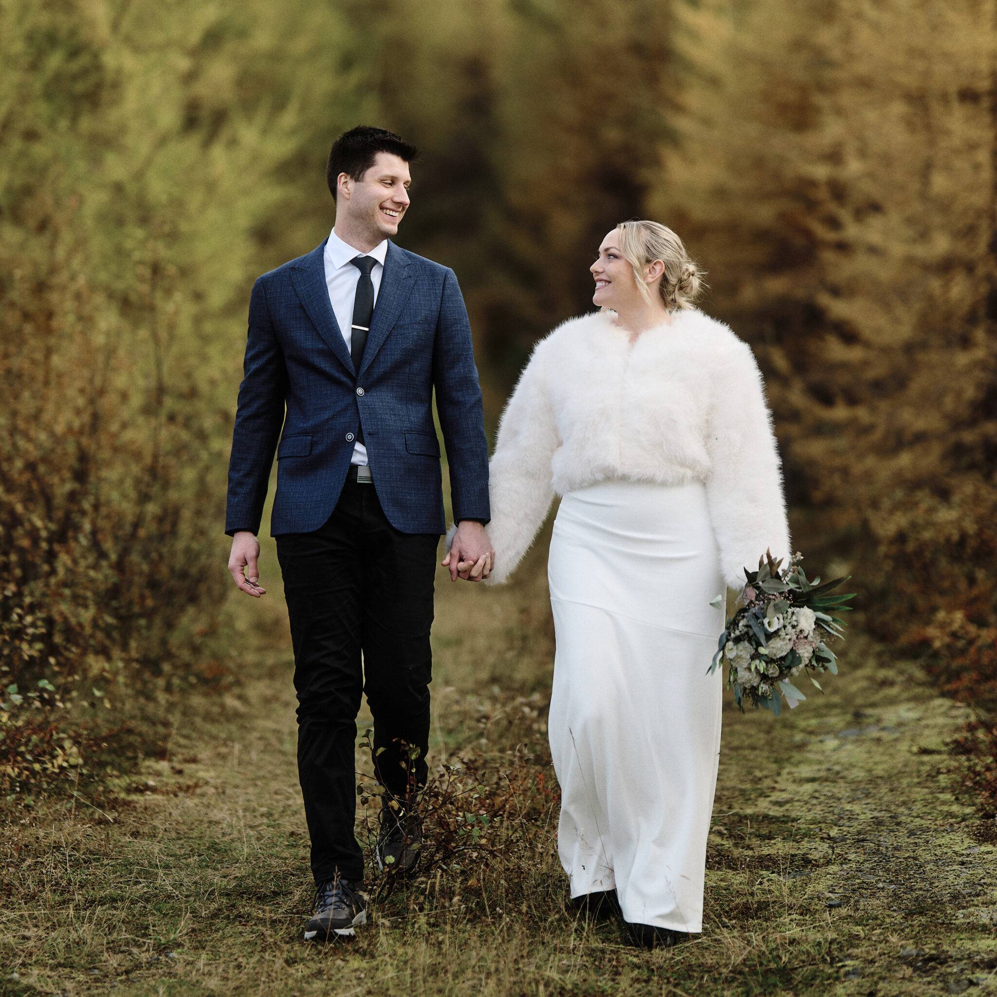 Romantic couple walking hand in hand through autumn forest trail during intimate outdoor wedding.