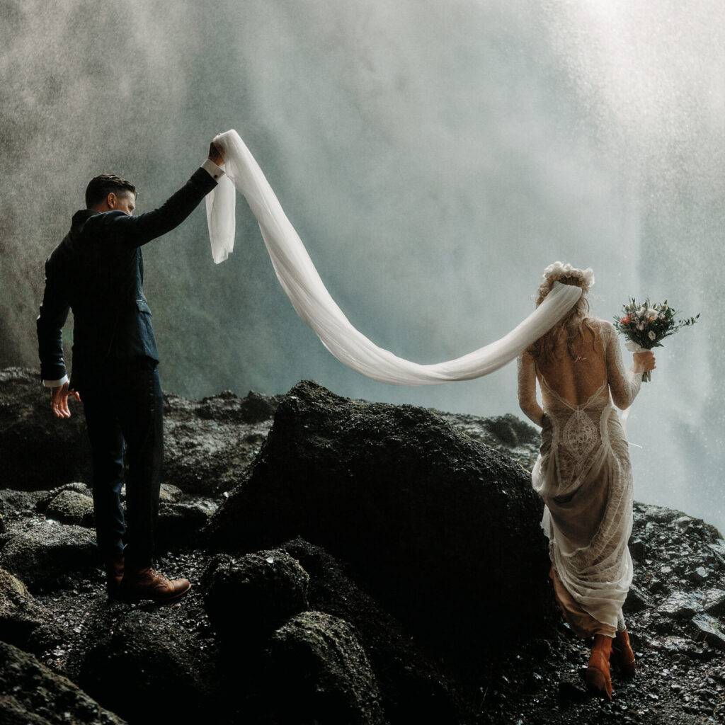 Romantic couple in wedding attire embracing beside misty icelandic waterfall and rugged volcanic rocks.