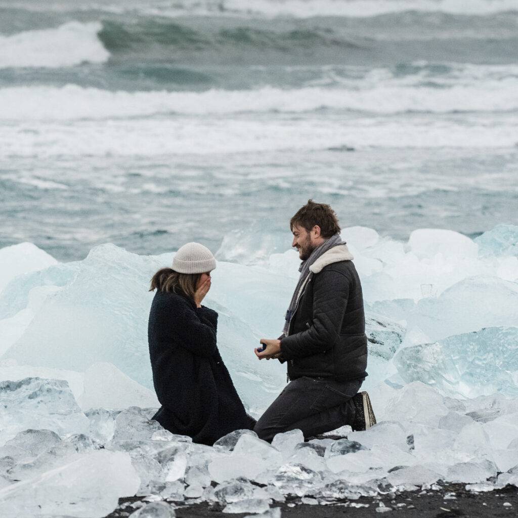 Man proposes to emotional woman on icy iceland shore with blue ice and crashing waves.