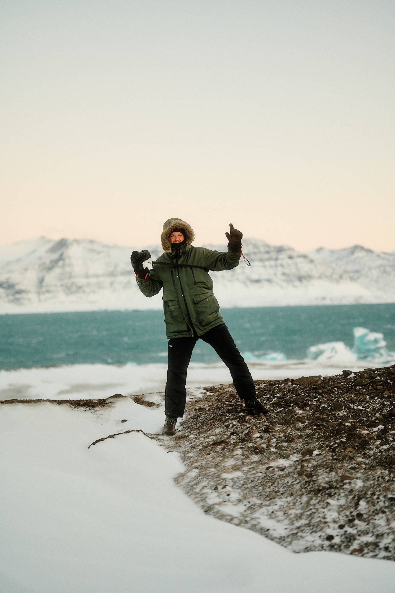 Person in winter coat celebrating near icy fjord with snow-covered mountains in iceland.