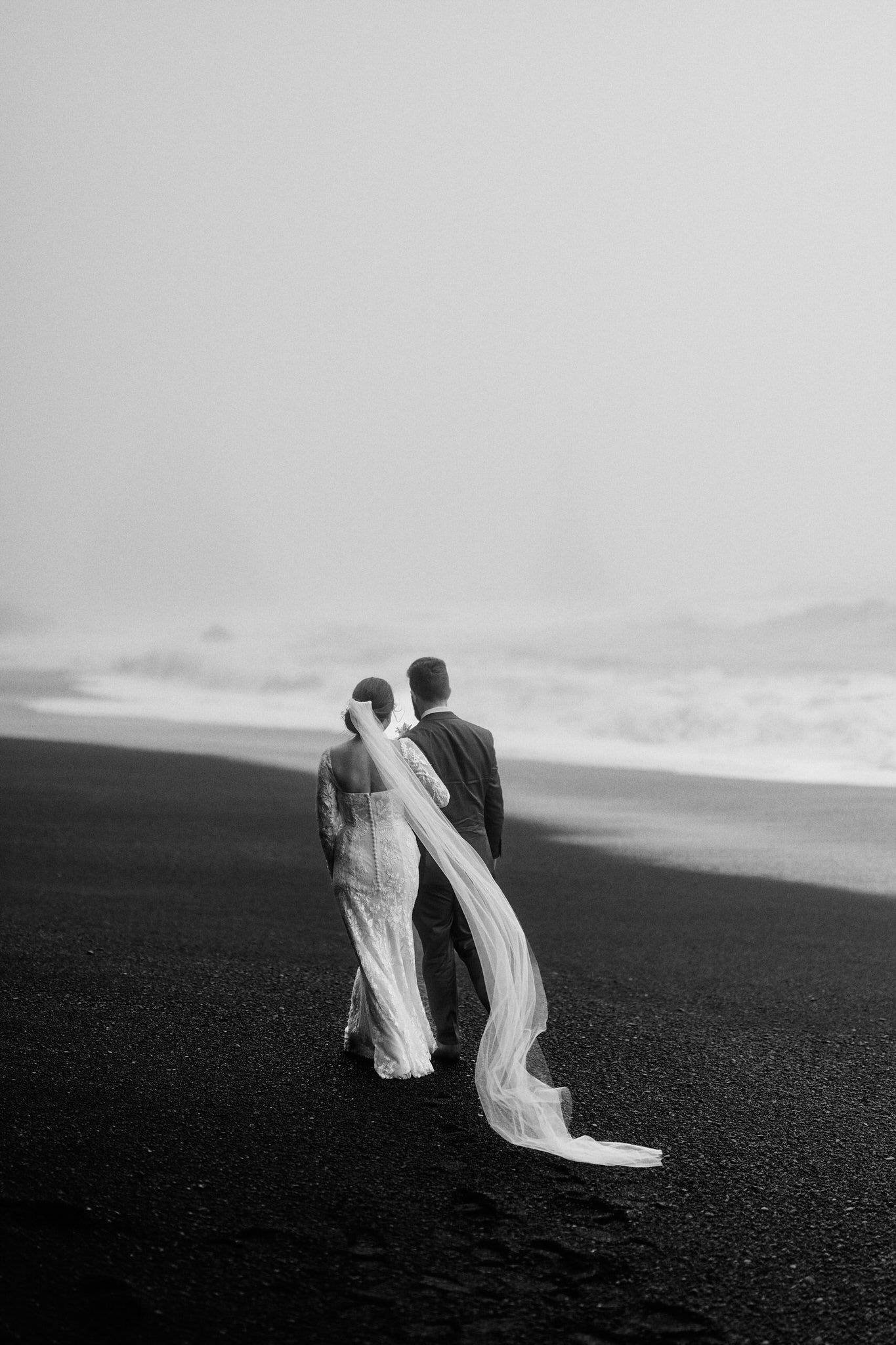 Newlywed couple walking on iceland’s black sand beach, showcasing love and natural beauty.